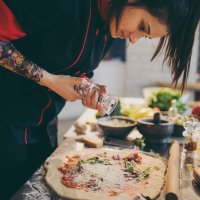 Foto. Tatoveret kok krydrer pizza i restaurantkøkken.