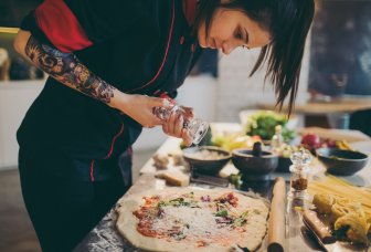 Foto. Tatoveret kok krydrer pizza i restaurantkøkken.